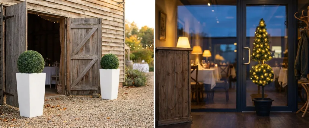 A pair of artificial boxwood balls in tall white planters at the open doors of a barn wedding venue, and a lighted artificial pine topiary inside the glass entrance of a restaurant at dusk.