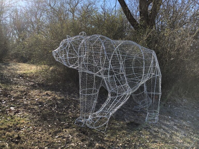 Bare wire-mesh armature in the shape of a walking bear, standing outdoors among leafless shrubs