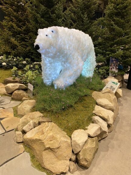 White lit bear sculpture on display among potted evergreens, an American flag visible in the background