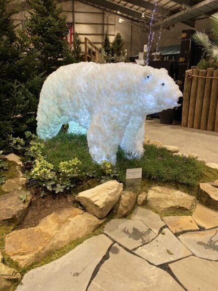 White lit bear sculpture on a grassy platform bordered by stones, in an indoor show setting