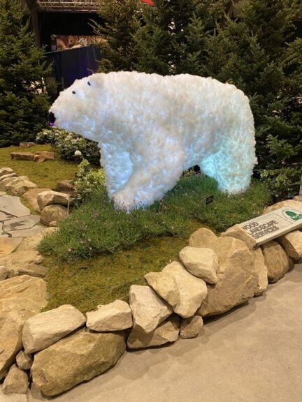 White, internally-lit bear sculpture on artificial turf, staged at a trade show with rock border and pine trees