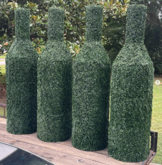 Four wine bottle shapes wrapped in green clipped foliage standing on an outdoor table