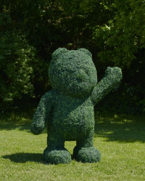 A standing green topiary bear with one arm raised, set against trees on a lawn