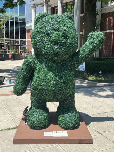 A standing waving bear topiary on a wooden base with a small branded sign, in a plaza setting