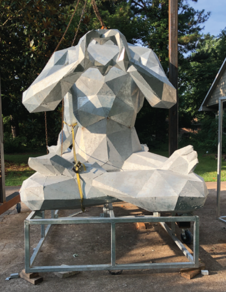 Galvanized steel figure sculpture suspended from a hoist, hands forming a heart shape