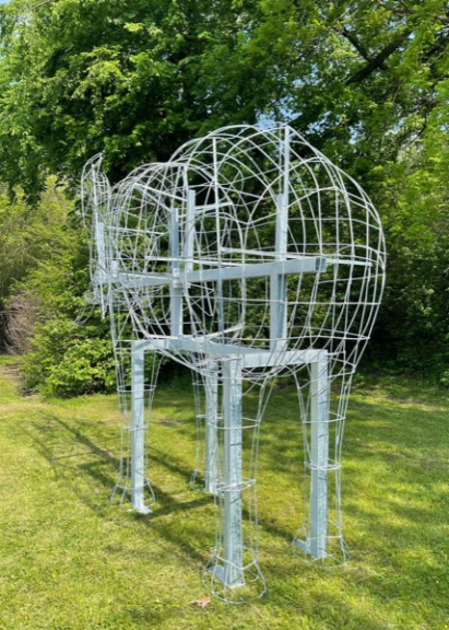 Unfoliaged wire elephant frame with a rounded, mesh-covered head section, on grass among trees.