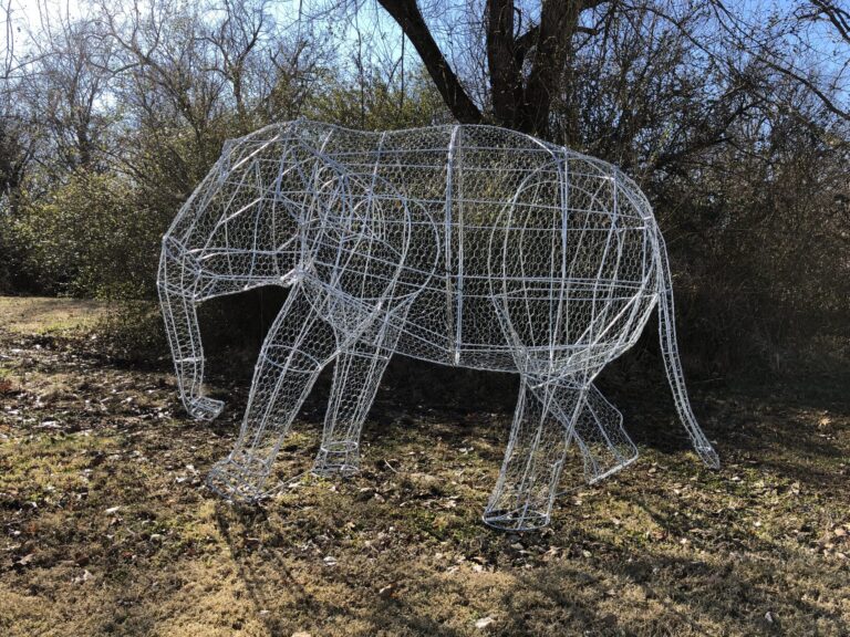 Silver wire-mesh elephant frame in a walking pose, standing outdoors among bare trees.