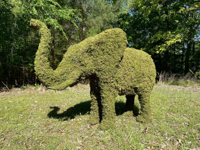 Moss-covered elephant topiary with trunk raised, standing in grass near a wooded tree line.