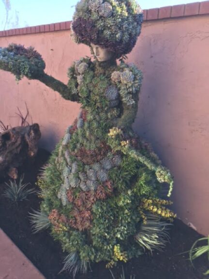 Full figure in a succulent dress reaching one arm toward a red flowering plant.