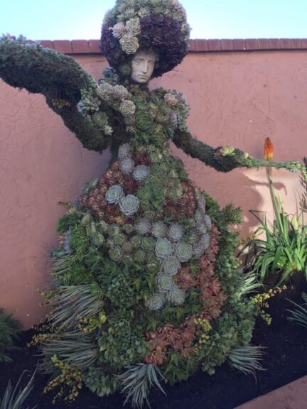 Full figure of a woman in a succulent-covered dress, arms outstretched, in a garden bed.