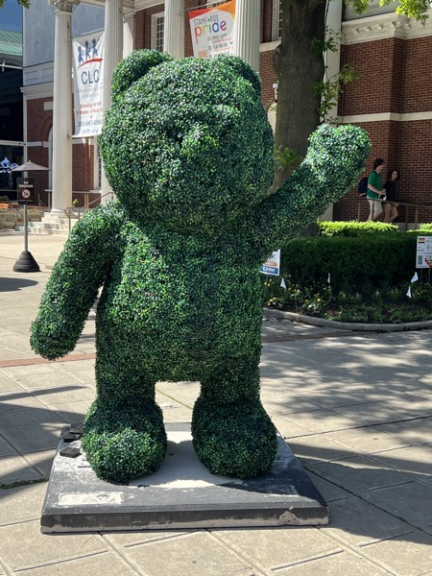 A standing waving bear topiary on a stone base outside a building, with event banners visible