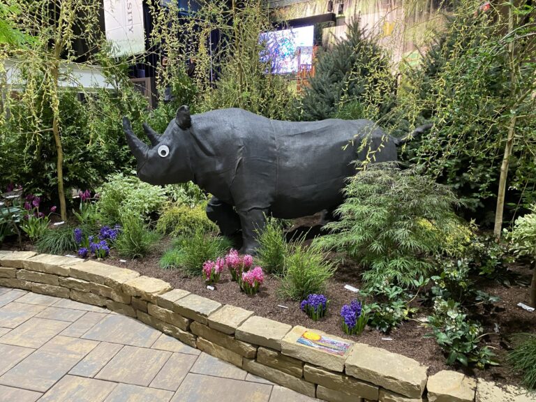 Black fabric rhino figure in a garden-show display with a stone wall, walkway, and banners behind it
