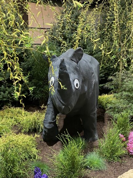 Close front view of a black fabric rhino head with two large googly eyes, willow branches overhead