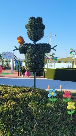 A boxwood scarecrow topiary installed at an outdoor park, with children's cartoon signage and figures in the background.