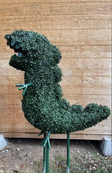 Side view of the cartoon-styled T-Rex topiary on green steel legs against a wood plank wall