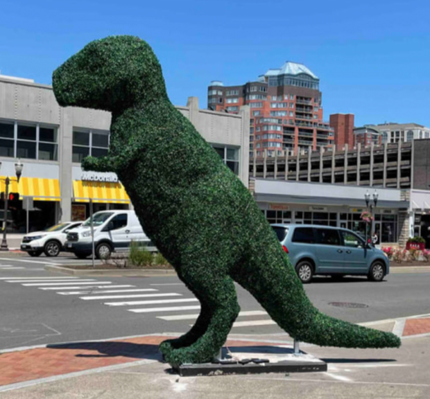 Tall green T-Rex topiary on a steel base plate at a city crossing, standing higher than the parked cars behind it