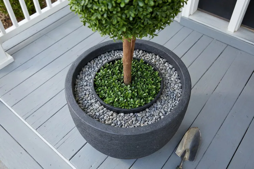 Looking down into a charcoal planter on a porch: the black grower's pot of an artificial topiary sits in the center with gray pea gravel packed around it almost to the rim.