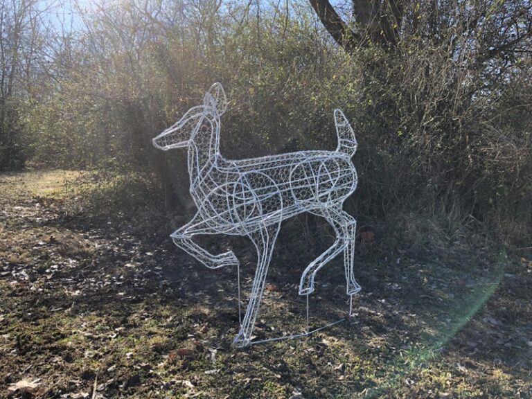 Mesh-wrapped doe frame in a mid-stride pose, staked in a sunlit field beside a bare hedgerow