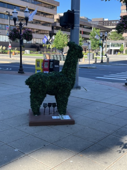 Green llama topiary on a plinth at a city intersection with a passing dog