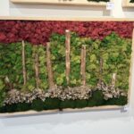 A framed wall panel with a red moss canopy above a green base, wood posts standing upright, and light pebbles scattered near the bottom.