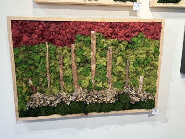 A framed wall panel with a red moss canopy above a green base, wood posts standing upright, and light pebbles scattered near the bottom.