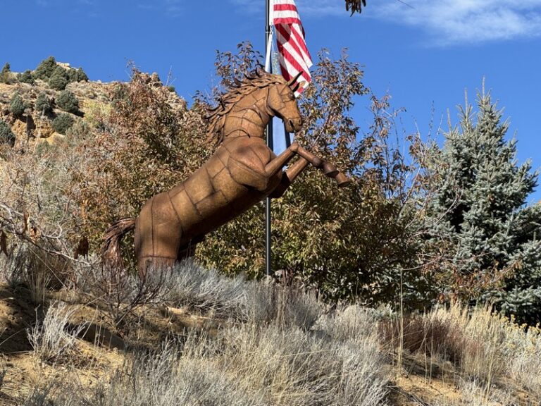 Rusted metal horse sculpture rearing on hind legs beside a flagpole and trees on a dry hillside