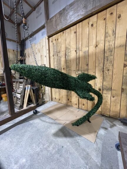 Green foliage-covered rocket topiary hanging diagonally from a chain hoist, tapered nose and small fin visible