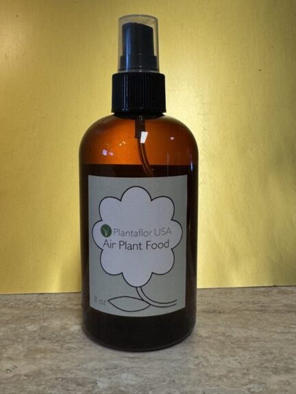 Amber glass spray bottle with a black pump mister and a label reading Plantaflor USA Air Plant Food, on a stone counter