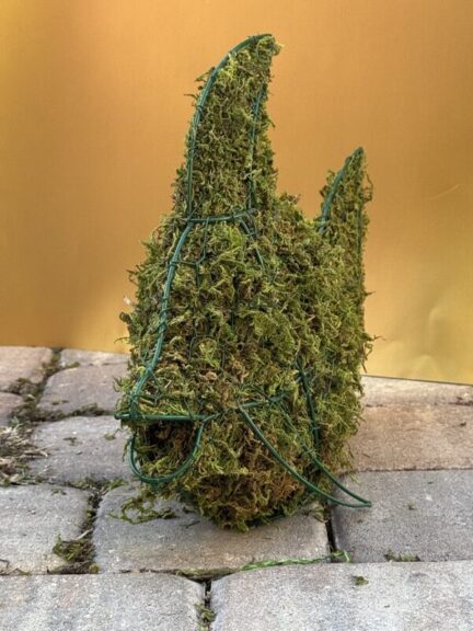 Moss-covered wire-frame fish topiary standing on pavers, open mouth and raised tail fin, gold backdrop