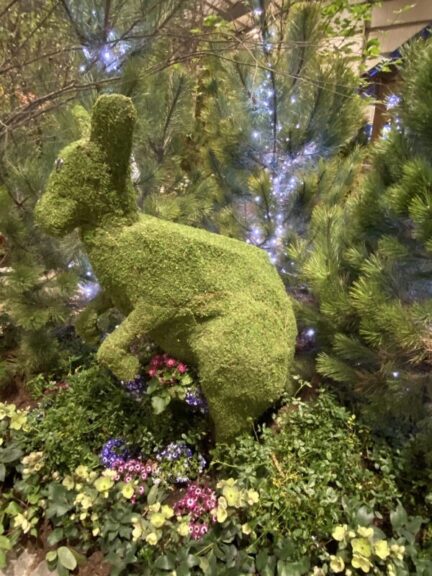 Side view of a moss kangaroo topiary among flowering plants and string-lit branches indoors