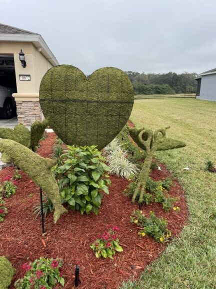 Moss scissors topiary on a garden stake, beside a large moss heart shape in a mulched bed