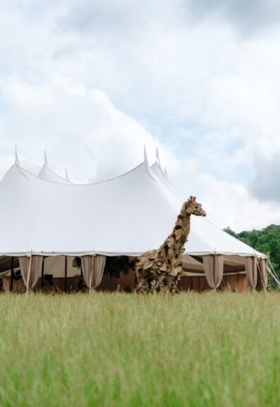 Foliage-covered giraffe sculpture standing in a grass field beside a large white event tent.