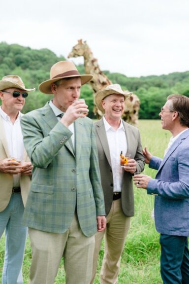 Group of men in blazers holding drinks, with the foliage-covered giraffe sculpture visible behind them.
