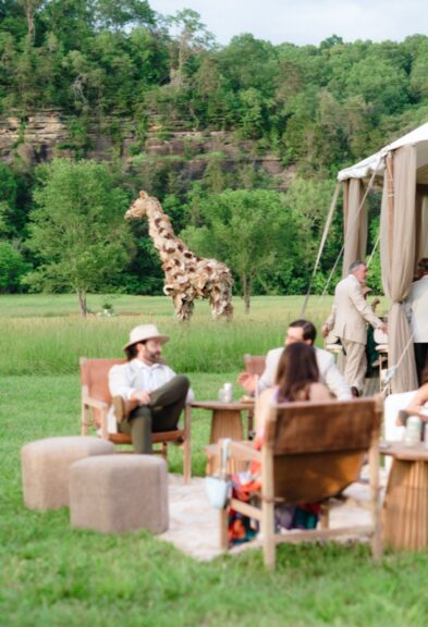 Giraffe sculpture in a grassy field near wooded cliffs, with an event tent and seated guests in the foreground.