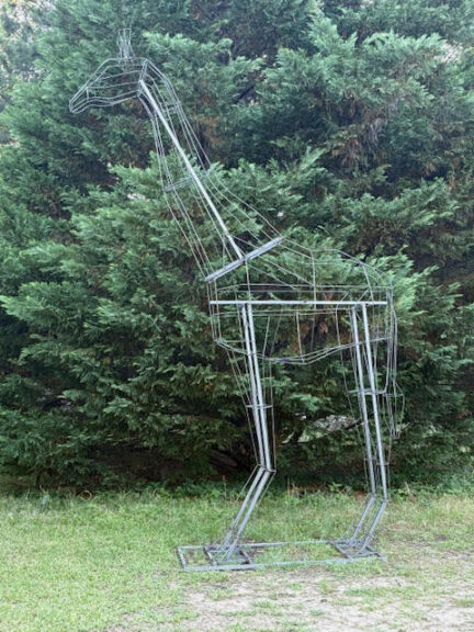 Bare wire-and-metal giraffe frame standing on grass in front of an evergreen hedge, unfinished.