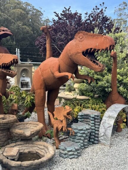 Rust-toned metal T-Rex sculpture with open jaws, beside a long-necked dinosaur and a smaller dragon-like figure