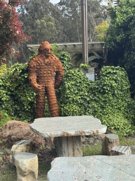 Rust-toned metal Bigfoot sculpture with a shingled, layered surface standing among garden hedges