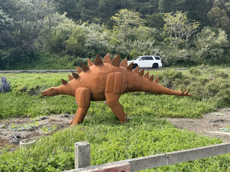 Rust-toned metal stegosaurus sculpture with spiked plates, standing in grass beside a road