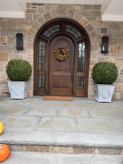 Boxwood ball topiary in a planter beside a wood and stone entry door.