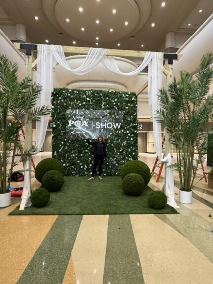A woman standing in front of a flower-covered green wall display, one hand raised toward the sign.