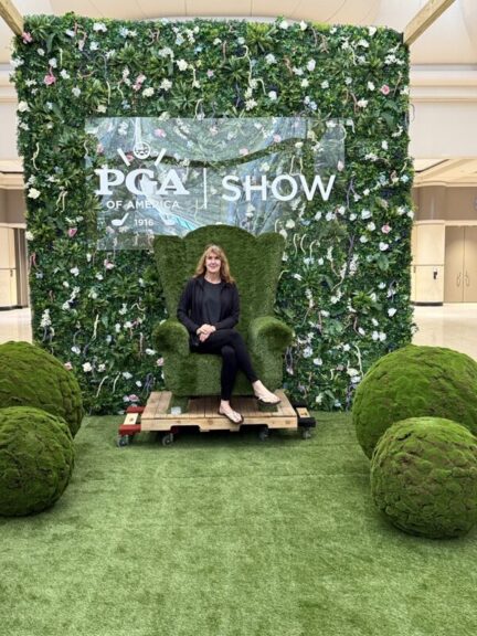 A woman seated in a turf-covered armchair in front of a finished flower-covered green wall and sign.