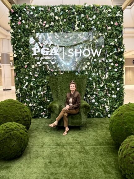 A woman seated in a turf-covered armchair in front of a flower-covered green wall and event sign.