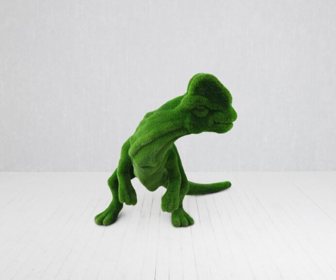 Dinosaur-shaped sculpture covered in green artificial turf, shown in a plain white room.