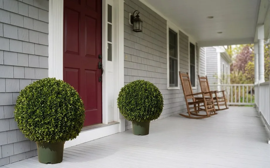A pair of artificial boxwood balls in green pots under the roof of a covered porch by a burgundy front door, with rocking chairs beyond.