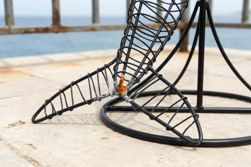 Close-up of the base of a black powder-coated wire topiary frame on a stone terrace by the sea, with the coating chipped at one joint and a small patch of orange rust on the bare steel.