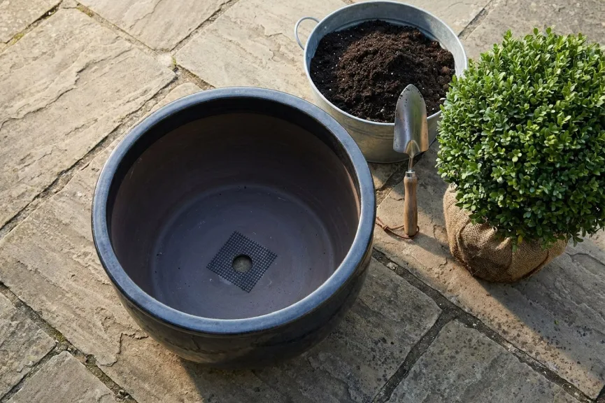 An empty dark glazed planter on a flagstone patio seen from above, a square of black mesh over the single drainage hole and no gravel inside, with a tub of potting mix, a trowel and a boxwood ball on its burlap root ball beside it.