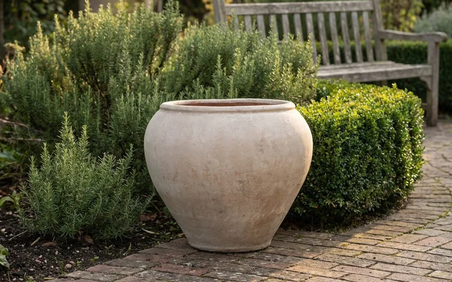 An empty wide-bellied stone-colored jar planter on a brick garden terrace beside rosemary and a clipped boxwood hedge.