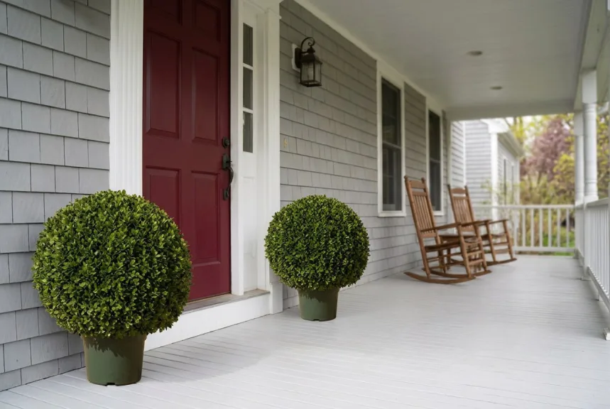 Two potted artificial boxwood balls beside a red front door on a covered porch