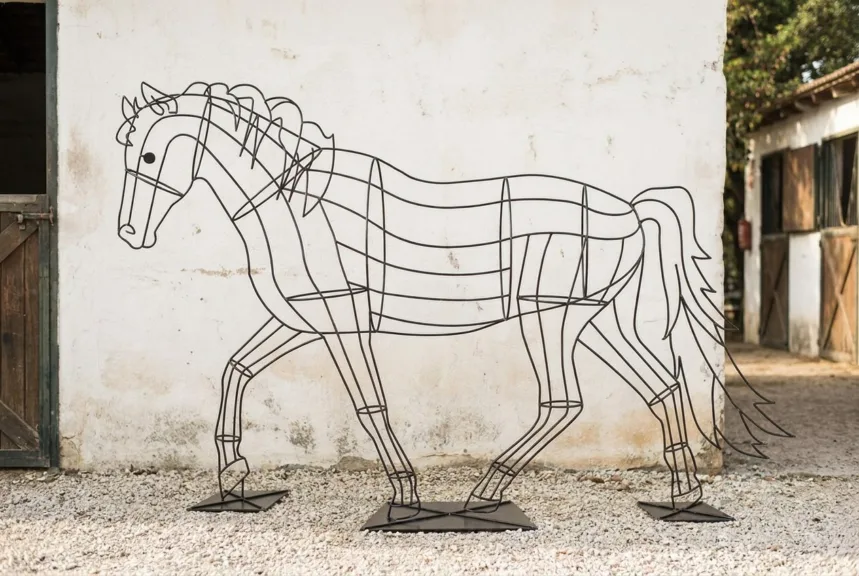 A bare welded steel horse topiary frame on a gravel yard in front of a whitewashed stable wall