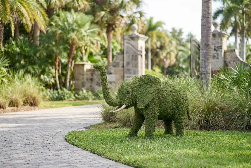 A five-foot moss elephant topiary with raised trunk on a lawn beside a zoo entrance path